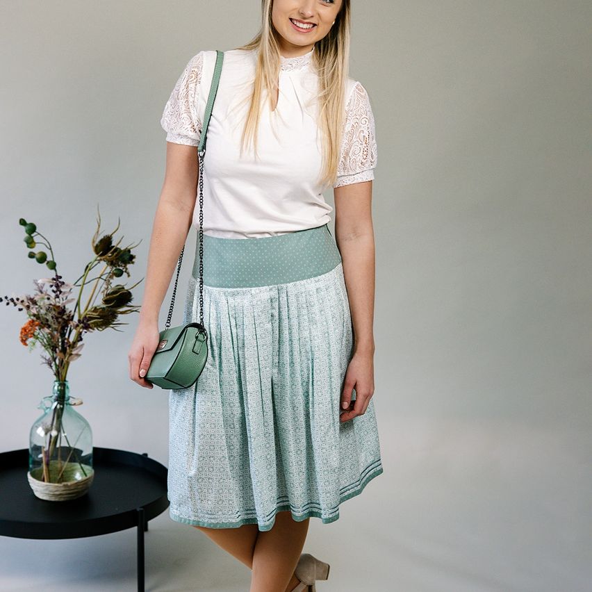 Eine Frau mit blonden Haaren steht vor einer weißen Wand und trägt ein weißes Oberteil mit einem grünen Gürtel, einem mintgrünen Faltenrock und einer grünen Umhängetasche. Ein Glasvase mit Blumen steht auf einem schwarzen Tisch hinter ihr.