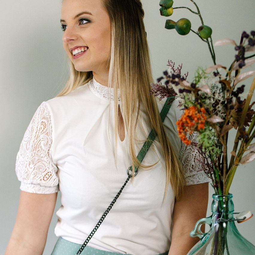 Eine Frau mit blonden Haaren lächelt, trägt ein weißes Oberteil mit Spitzenärmeln und einen grünen Riemen. Sie steht neben einer Vase mit einem Blumenarrangement.