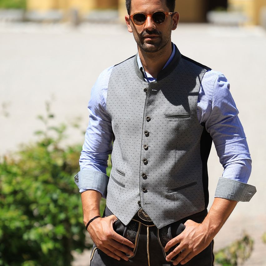 Ein Mann in einem blauen Hemd und grauer Weste steht draußen mit Sonnenbrille, Händen in den Taschen und einem Gürtel.