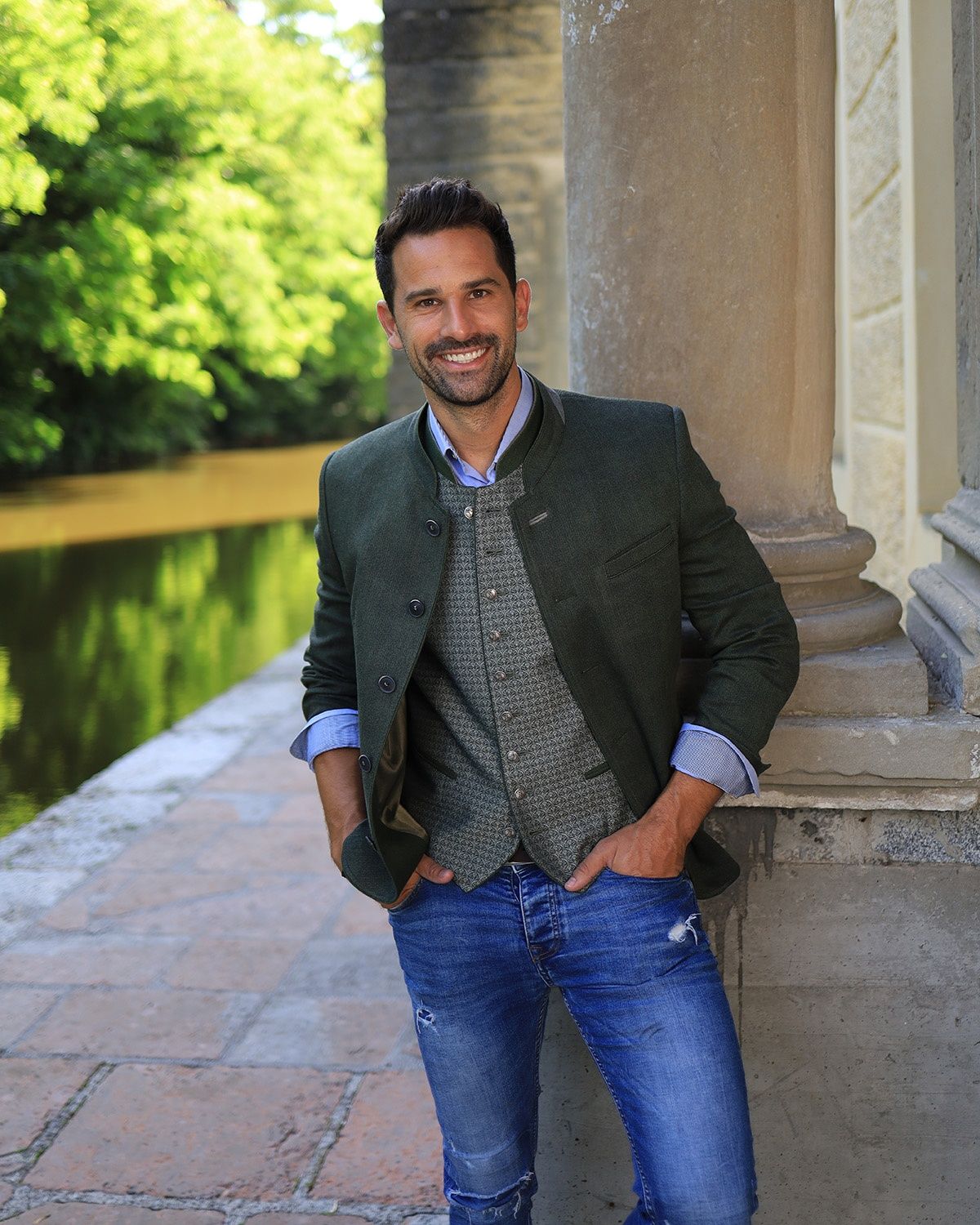 Ein Mann steht lächelnd und posiert für ein Foto, trägt einen grünen Blazer über einem gemusterten Weste und blaue Jeans, mit einer großen Säule hinter ihm.