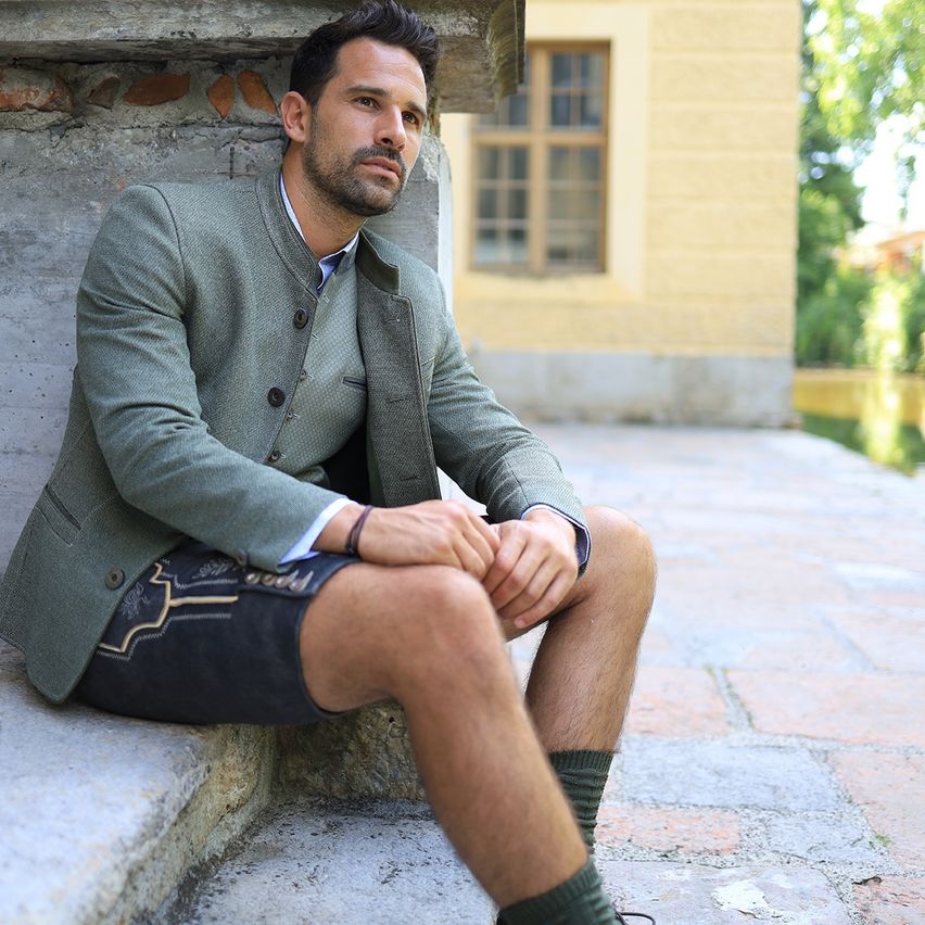 Ein Mann in einem grünen Blazer, schwarzen Shorts und grünen Socken sitzt auf einer Steintreppe. Er schaut nach links. Hinter ihm steht ein gelbes Gebäude mit Fenstern und Bäumen.