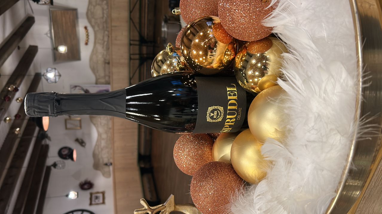 Eine Flasche Sprudel Edel Wein liegt auf einem Haufen goldener und brauner glitzernder Weihnachtskugeln. Das Etikett lautet 'Sprudel Edel Deluxe'.