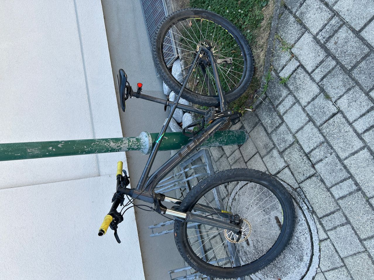Ein schwarzes Mountainbike liegt auf dem Kopf auf einem gepflasterten Gehweg neben einem grünen Pfosten. Das Vorderrad ist auf dem Boden und das Hinterrad in der Luft.