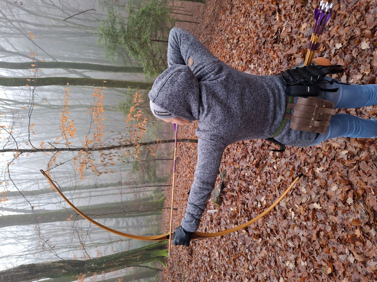 Eine Person in einer grauen Kapuzenjacke liegt auf dem Boden und hält einen Bogen und einen Pfeil. Der Bogen und der Pfeil sind braun, und der Pfeil hat eine lila Feder. Die Person befindet sich in einem Wald mit trockenen Blättern auf dem Boden. Der Wald ist neblig.