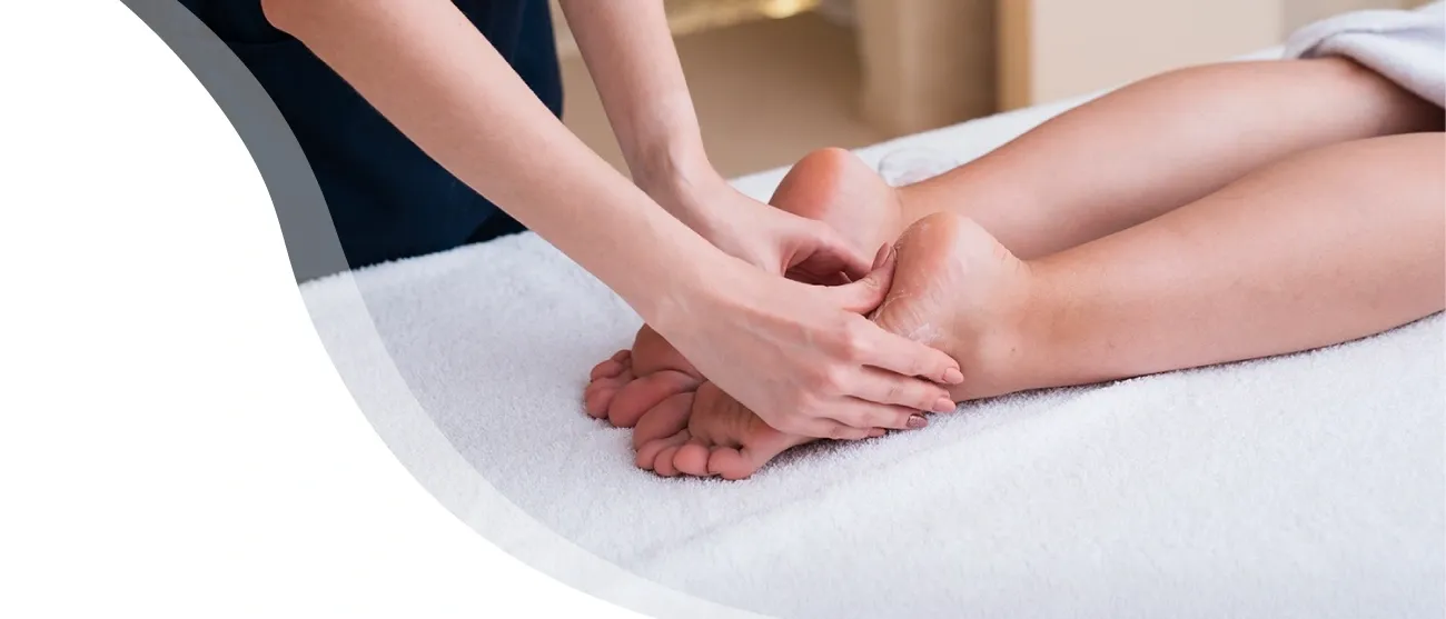 Eine Frau in einem blauen Kleid massiert die Füße einer Kundin, die auf einem weißen Handtuch auf einem Bett liegt.