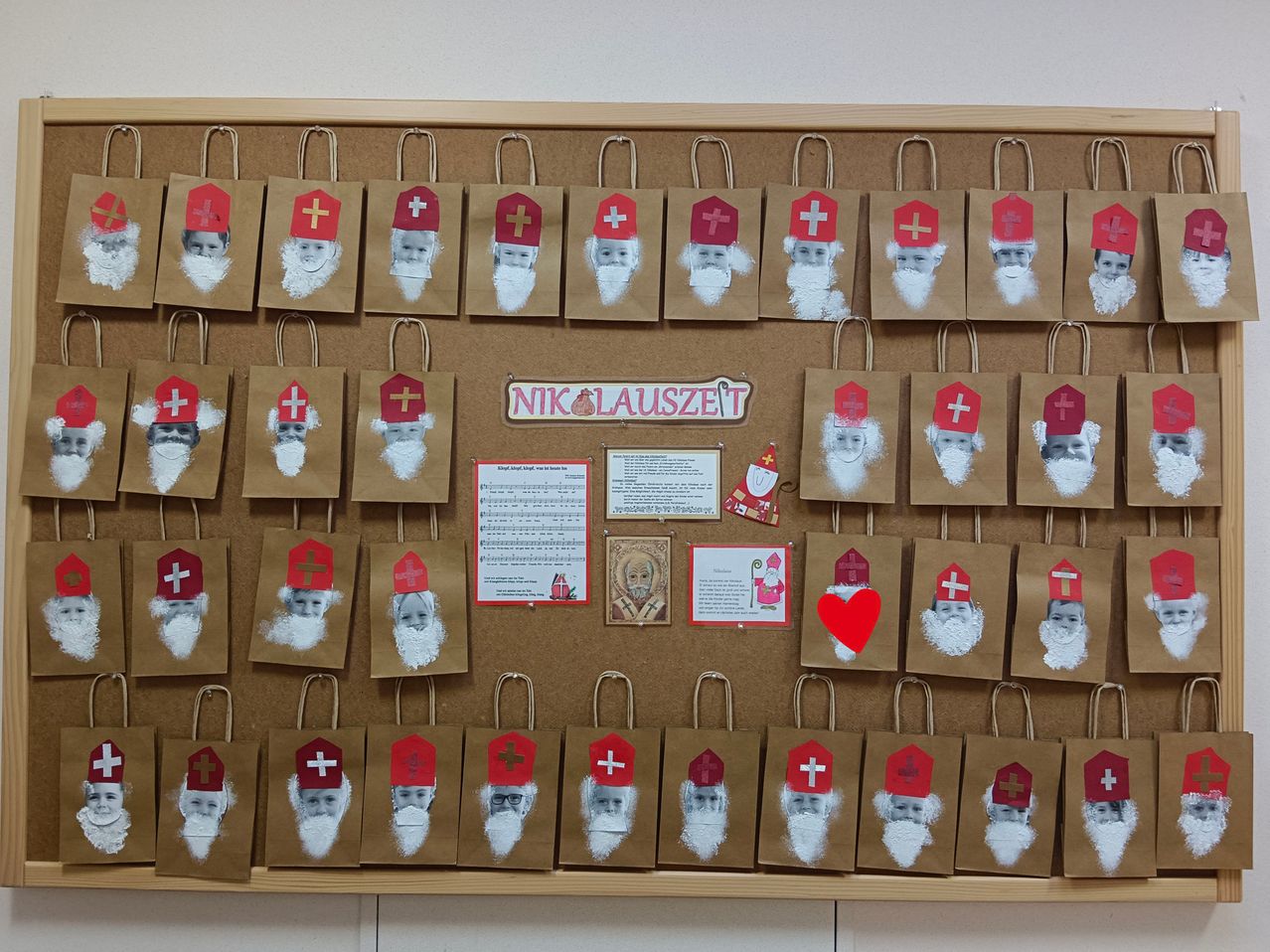 Ein Bulletin-Board zeigt zahlreiche braune Papiertüten, die mit weißen Bärten und roten Kreuzen verziert sind, wahrscheinlich für eine Sankt-Nikolaus-Feier. Ein rotes Herz und verschiedene Karten schmücken das Board.