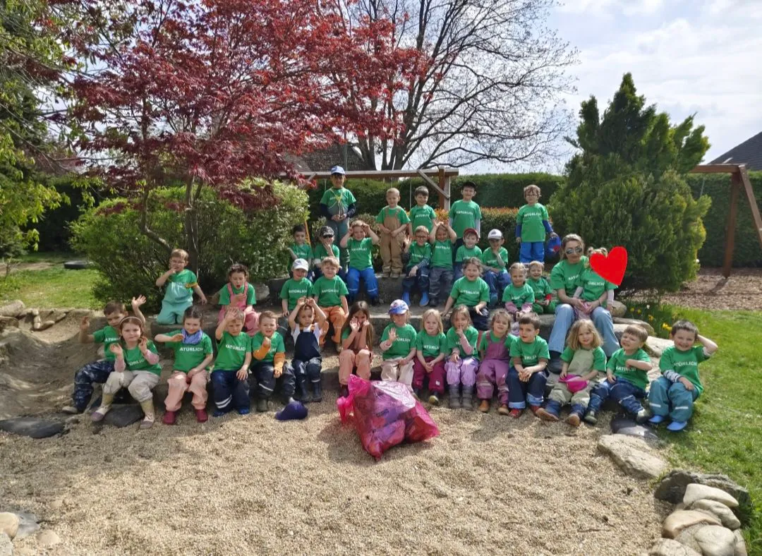 Eine Gruppe von Kindern in grünen T-Shirts und Latzhosen posiert für ein Foto im Freien. Sie sind von Bäumen und Büschen umgeben. Einige Kinder tragen Mützen. Ein rotes Herz ist auf der linken Seite angeheftet.