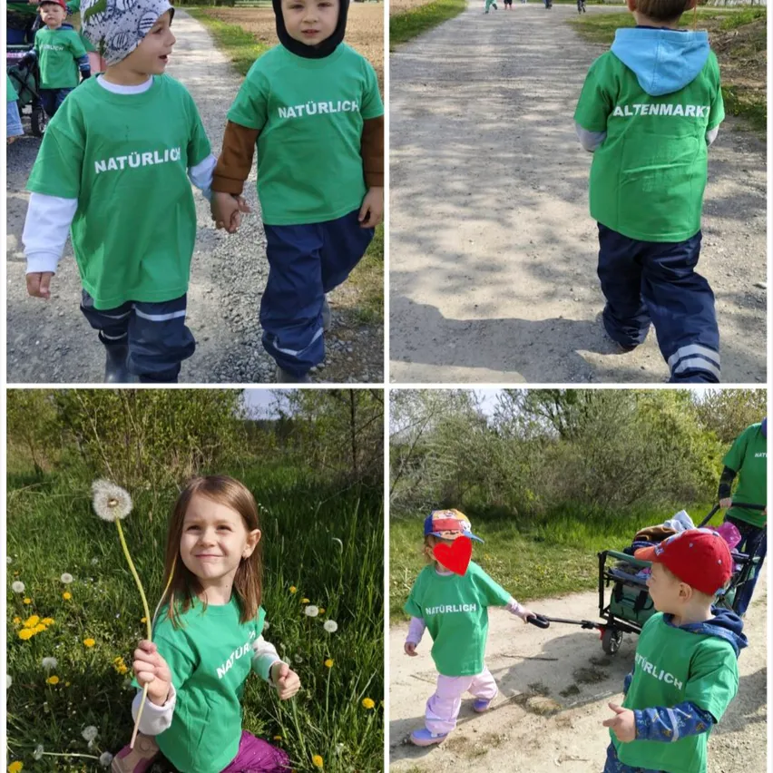 Mehrere Kinder tragen grüne 'Naturlich'-Shirts, manche halten Hände, andere gehen mit einem Kinderwagen, eines hält eine Pusteblume, in einem Park mit Schotterwegen.