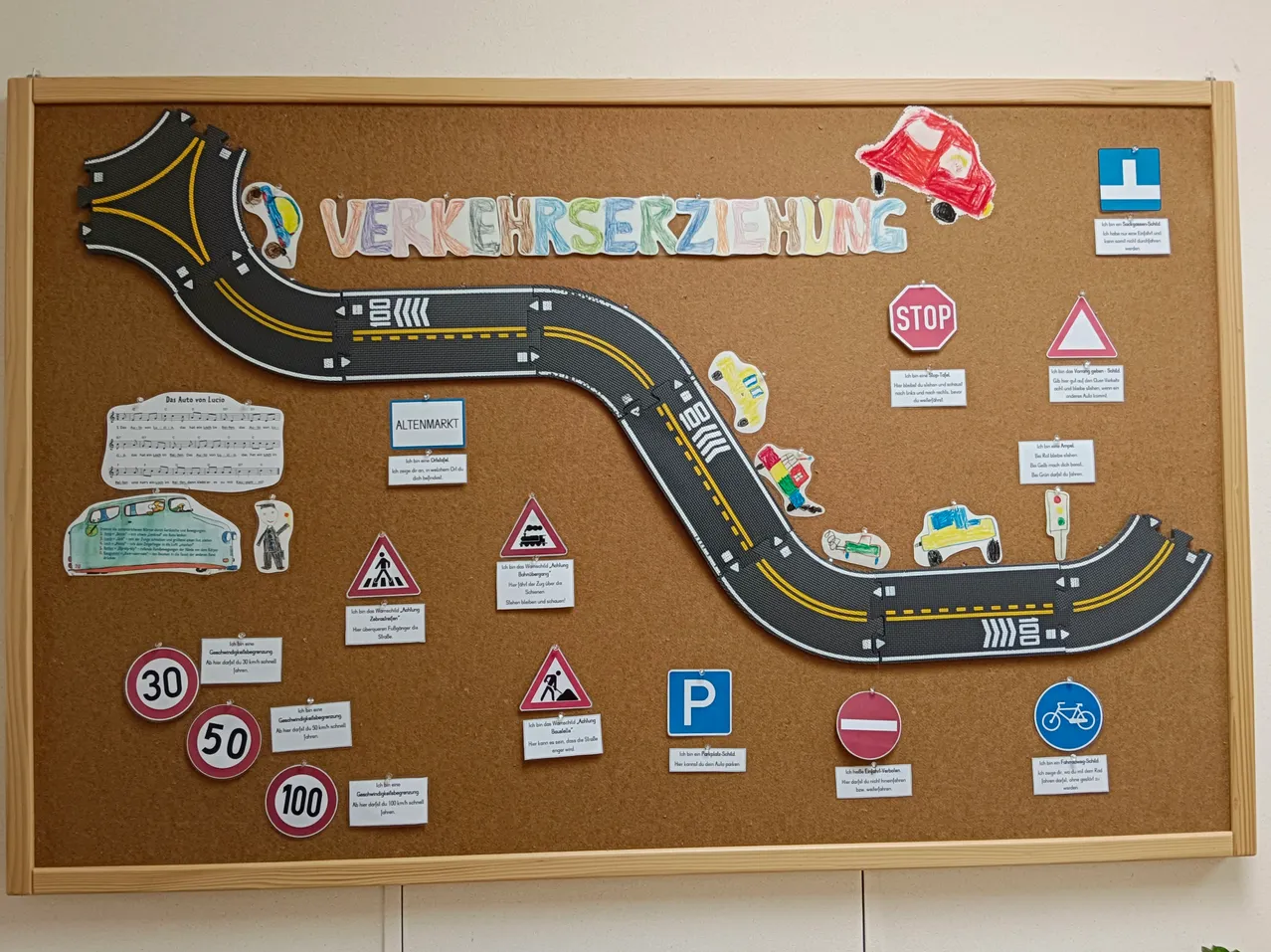 Ein Korkbrett mit einer gezeichneten Straße, dem Wort 'Verkehrszeichnung' und verschiedenen Verkehrszeichen wie 'Stop', 'P' und Geschwindigkeitsbegrenzungsschildern. Es gibt auch einige kleine Ausschnitte von Autos und Fußgängern.