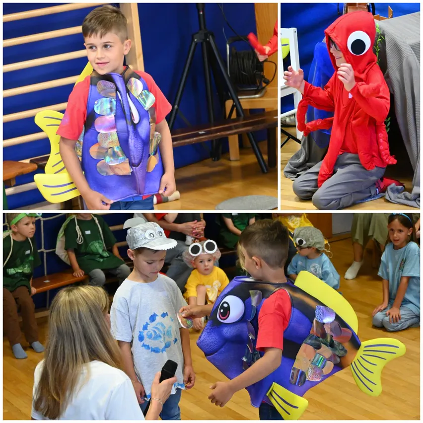 Eine Gruppe von Kindern in Meeres-Themen-Kostümen. Ein Kind in einem Fisch-Kostüm, ein anderes in einem Krebs-Kostüm und andere beobachten. Die Umgebung scheint ein Klassenzimmer oder ein ähnliches Innenumfeld zu sein.