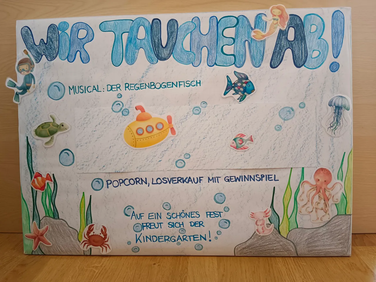 Ein Unterwasser-Poster mit dem Titel 'Wir tauchen ab' in blauen Buchstaben. Es zeigt ein U-Boot, Fische und Meerestiere. Es steht 'Musical: Der Regenbogenfisch,' 'Popcorn, Losverkauf mit Gewinnspiel,' und 'Freue dich auf ein schönes Fest im Kindergarten!'