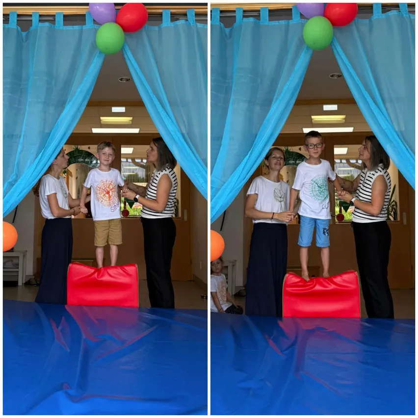Zwei Frauen helfen zwei Jungen, auf roten Kissen in einem Raum mit blauen Vorhängen und Ballons zu stehen.