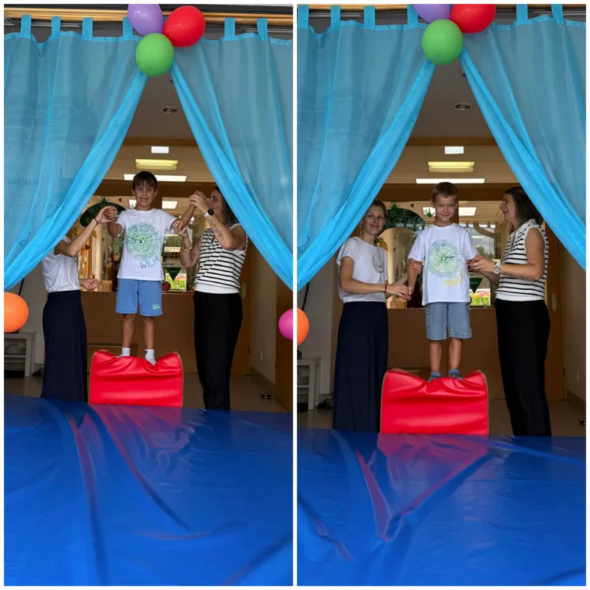 Zwei junge Jungen stehen auf roten Kissen auf einem blauen Teppich, wobei Erwachsene ihre Hände halten. Bunte Luftballons sind an den blauen Vorhängen hinter ihnen befestigt.
