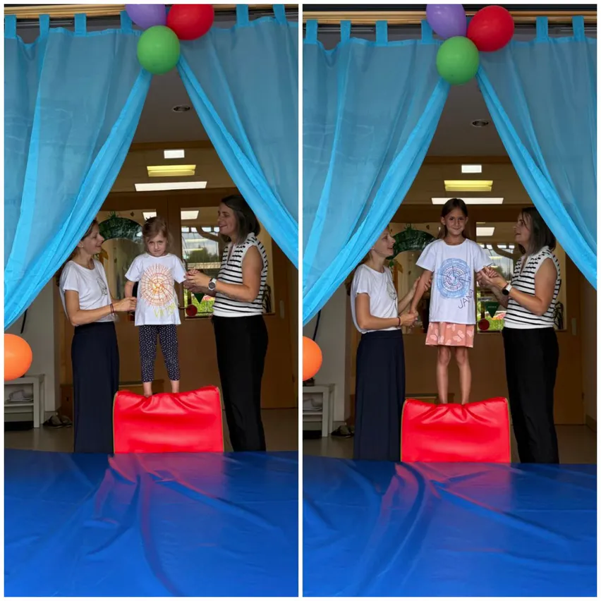 Zwei Frauen und ein junges Mädchen stehen auf einem roten Kissen auf einem blauen Teppich. Sie lächeln und scheinen für ein Foto zu posieren. Der Raum hat blaue Vorhänge und Luftballons.