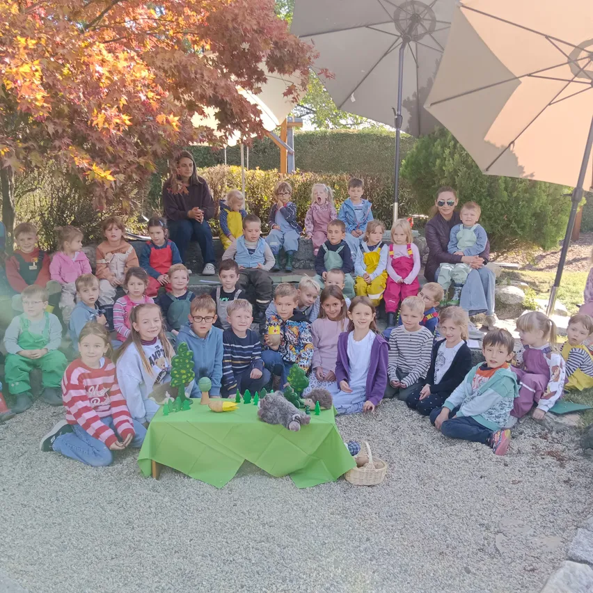 Eine Gruppe von Kindern und Erwachsenen versammelt sich in einem Garten. Einige sitzen auf dem Boden, andere stehen. Ein Tisch mit einem grünen Tuch steht in der Mitte, und einige Spielzeuge sind darauf. Die Erwachsenen sitzen auf einer Bank unter einem Regenschirm.