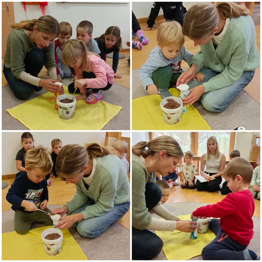 Eine Gruppe von Kindern und Erwachsenen sitzt auf dem Boden und ist mit einer Aktivität beschäftigt. Jedes Kind hält einen Becher und einen Löffel und gießt etwas in einen Eimer. Die Erwachsenen leiten die Kinder.