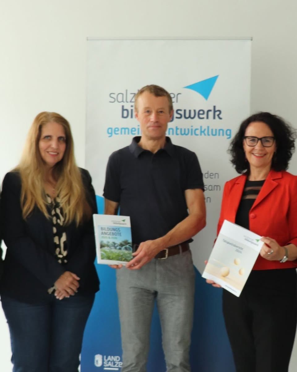 Drei Personen stehen vor einem Banner und halten Dokumente in den Händen. Der Mann in der Mitte hält ein Dokument mit dem Titel 'Bauangebot'. Die Frau rechts hält ein Dokument mit dem Wort 'Wettbewerb'. Sie alle lächeln.