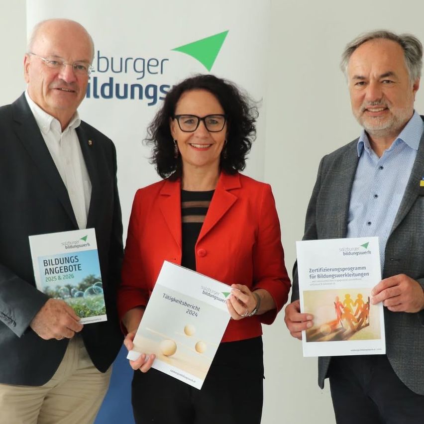 Drei Personen stehen zusammen und halten Bücher in den Händen. Der Mann links trägt eine Brille und einen Anzug. Die Frau in der Mitte trägt einen roten Blazer und eine Brille. Der Mann rechts trägt einen hellen Blazer und hat eine kleine Flagge am Revers.