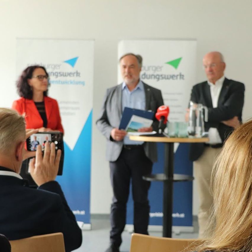 Eine Gruppe von Menschen steht vor einem Podium mit Mikrofonen. Ein Mann in einem grauen Anzug spricht. Hinter ihm hört eine Frau in einem roten Blazer zu. Ein weiterer Mann in einem schwarzen Anzug steht neben ihm. Eine Person im Vordergrund macht ein Foto.