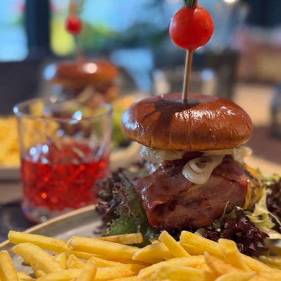 Ein Nahaufnahme eines Sandwiches mit Salat, Zwiebeln und einer Kirschtomate oben, begleitet von einem Getränk und Pommes.