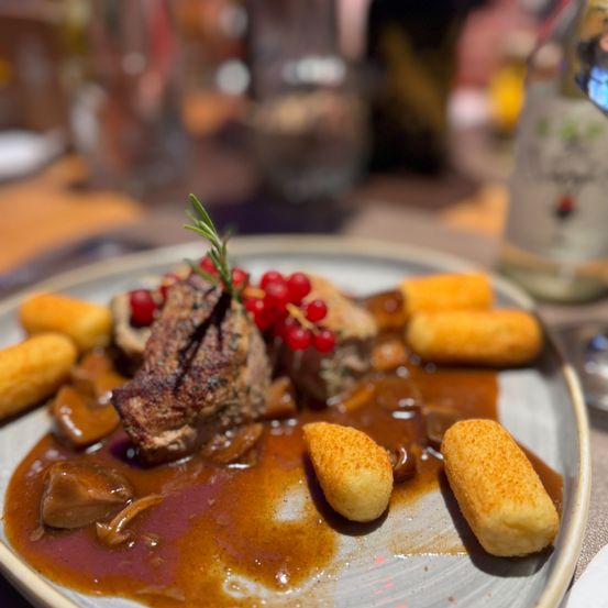 Ein Teller mit Rehmedallions in Rotbeerensauce, begleitet von gebratenen Champignons und kleinen goldenen Kroketten, garniert mit einem Rosmarinzweig.