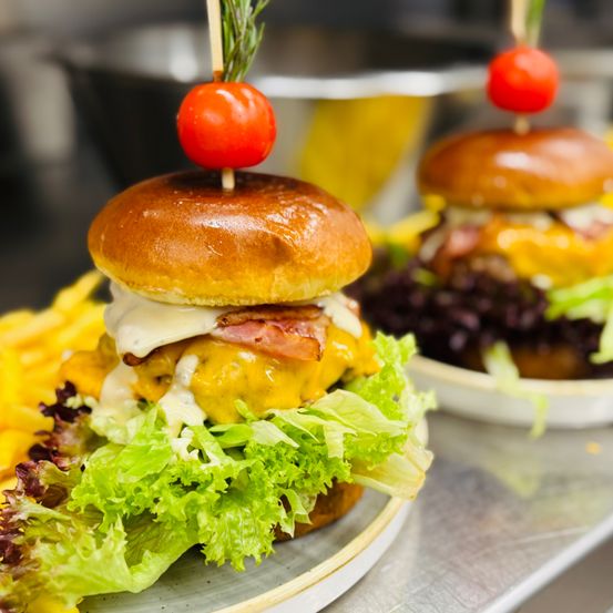 Eine Nahaufnahme von zwei Hamburgern mit Salat und Speck, gekrönt von einer Kirschtomate auf einem Teller. Ein weiterer Teller mit einem Hamburger im Hintergrund.