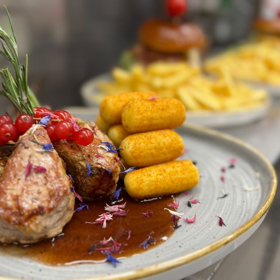 Ein Teller mit Steak in Sauce, garniert mit roten Beeren und Kräutern, begleitet von Kartoffelklößchen und Pommes auf einem Tablett.