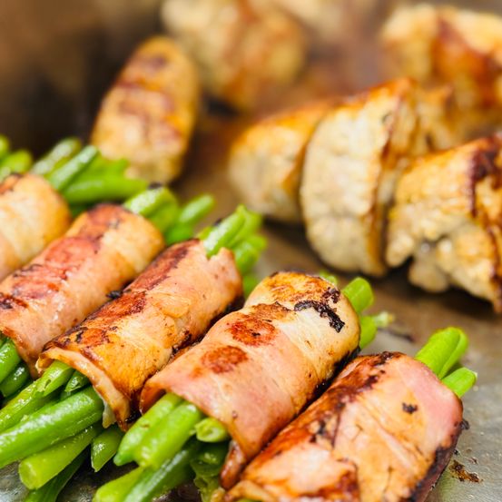 Gegrillte Hähnchenbrust, umwickelt mit Speck und grünen Bohnen auf einem Metalltablett, mit anderem gegrilltem Hähnchen im Hintergrund.