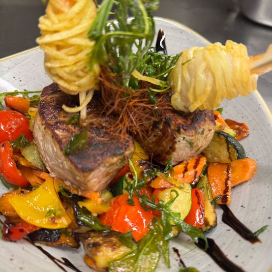 Ein Teller mit gegrilltem Steak, garniert mit frittierter Pasta, Kräutern und einer Mischung aus gegrilltem Gemüse.