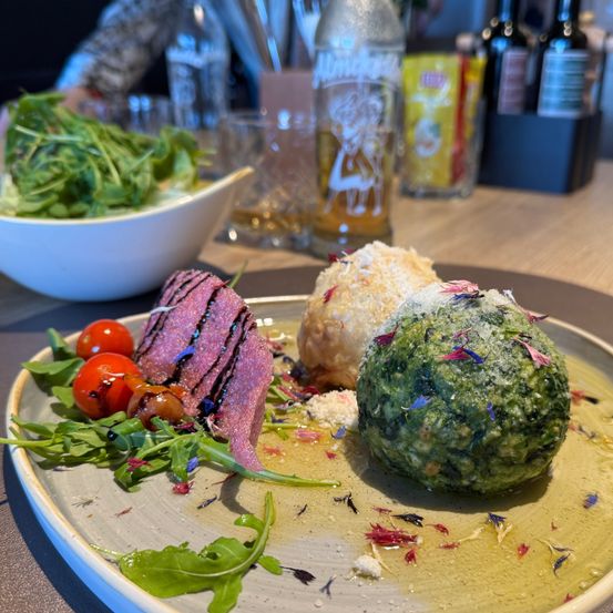Ein Teller mit Steak, grüner Kugel und Salat steht auf einem Holztisch mit Weinflaschen und einer Schüssel mit Salat im Hintergrund.
