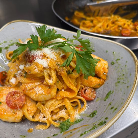 Ein Teller mit Garnelen-Pasta, garniert mit Petersilie und einer Käsestreuung. Im Hintergrund wird ein weiteres Gericht in einem Topf zubereitet.
