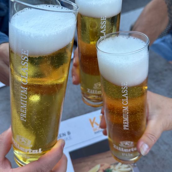 Drei Hände halten hohe Gläser mit Bier und schaumigen Köpfen. Die Gläser sind mit 'Premium Klasse' und 'Zillertal' bedruckt. Der Hintergrund zeigt ein teilweise sichtbares Menü.