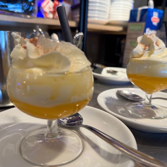 Zwei Dessertgläser mit Schlagsahne obenauf, auf weißen Tellern platziert. Neben jedem Glas befindet sich ein Löffel. Der Hintergrund zeigt eine verschwommene Ansicht eines Regals mit gestapelten Tellern.