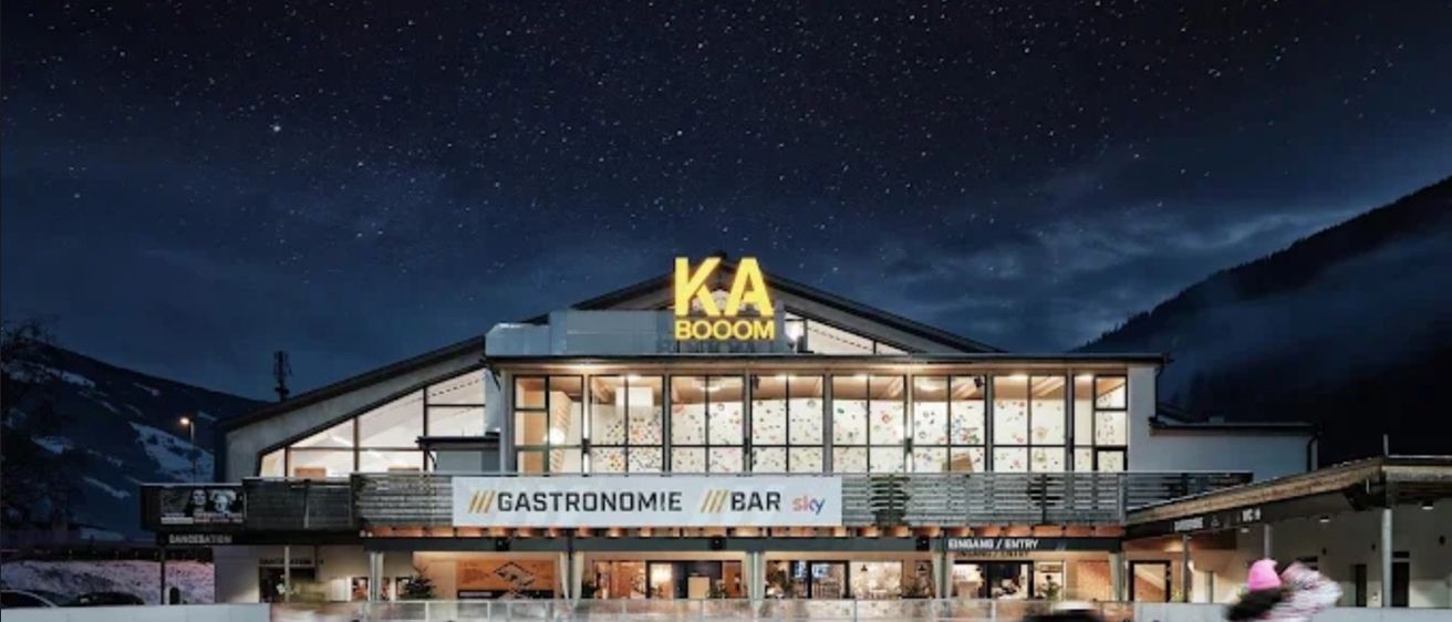 Eine Eisbahn mit Menschen, die unter einem großen Schild mit der Aufschrift KA BOOM Schlittschuh laufen. Die Bahn ist beleuchtet, und im Vordergrund steht eine Pinguinstatue.