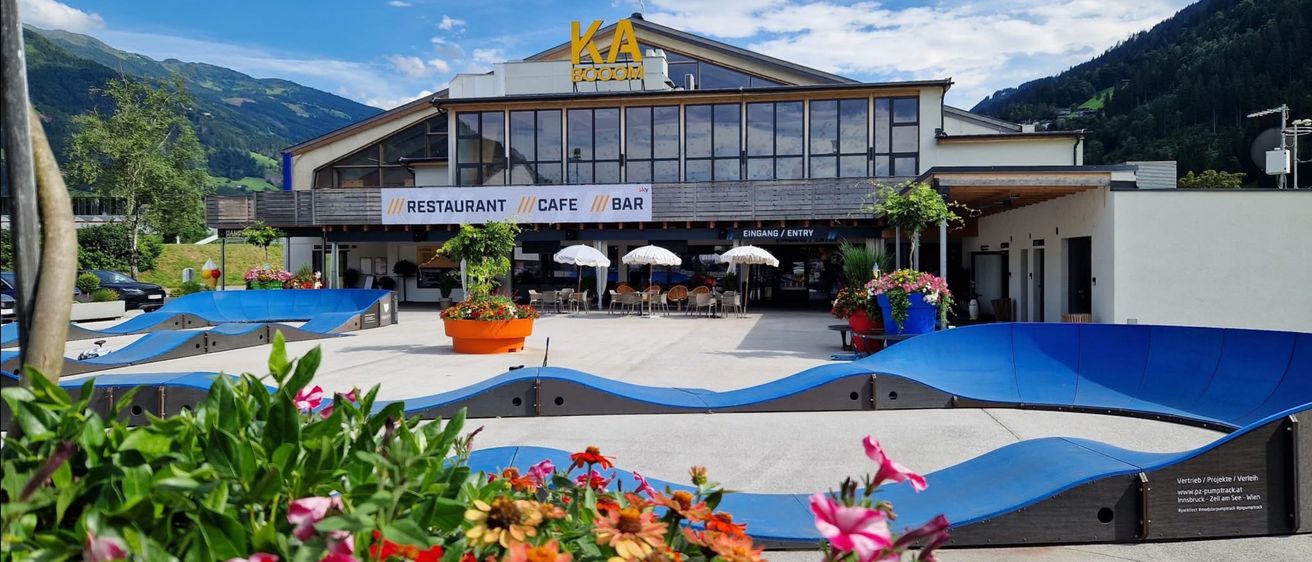 Ein modernes Gebäude mit einem großen Schild, das 'KA BOOM' zeigt, mit Restaurant, Café und Bar. Es ist von Bergen umgeben, und der Eingang hat draußen Schirme und Tische.