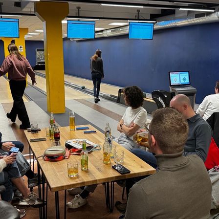 In einer Bowlingbahn versammeln sich mehrere Personen um einen Tisch, genießen Getränke und beobachten ein Spiel. Eine Frau läuft auf der Bowlingbahn.