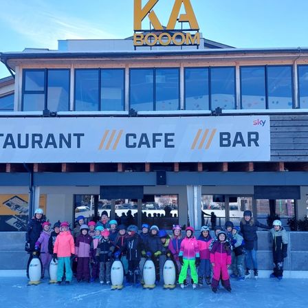 Eine Gruppe von Kindern in Winterkleidung und mit Eislaufschuhen posiert vor dem KA BOOM-Gebäude, das ein Schild für Restaurant, Café und Bar trägt.