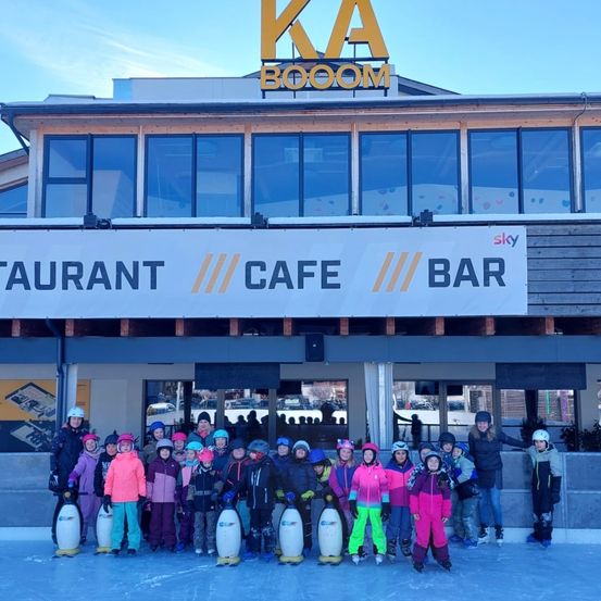Eine Gruppe von Kindern in Winterkleidung und mit Eislaufschuhen posiert vor dem KA BOOM-Gebäude, das ein Schild für Restaurant, Café und Bar trägt.