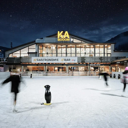 Eine Nachtszene bei KA BOOM, einer Eislaufbahn mit einem Gebäude, das Gastronomie und eine Bar anbietet, beleuchtet unter einem sternenklaren Himmel. Menschen schlittern um eine Pinguinstatue herum.