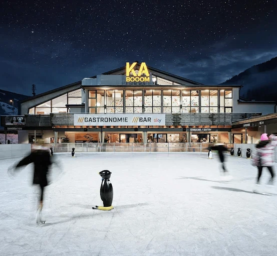 Eine Nachtszene bei KA BOOM, einer Eislaufbahn mit einem Gebäude, das Gastronomie und eine Bar anbietet, beleuchtet unter einem sternenklaren Himmel. Menschen schlittern um eine Pinguinstatue herum.