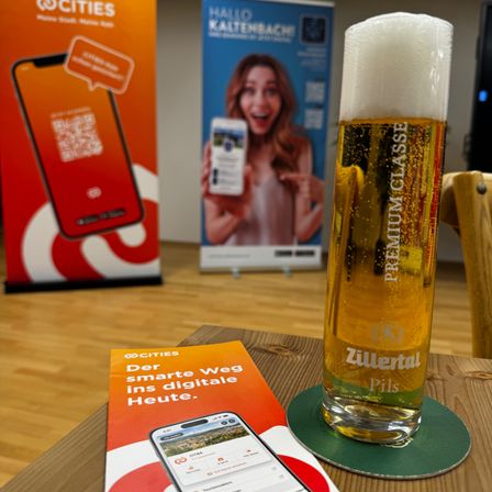 Ein Glas Zillertal Bier steht auf einem Untersetzer neben einem Prospekt und einem Smartphone auf einem Holztisch. Im Hintergrund befinden sich Banner mit Werbeinhalten.