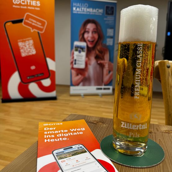 Ein Glas Zillertal Bier steht auf einem Untersetzer neben einem Prospekt und einem Smartphone auf einem Holztisch. Im Hintergrund befinden sich Banner mit Werbeinhalten.