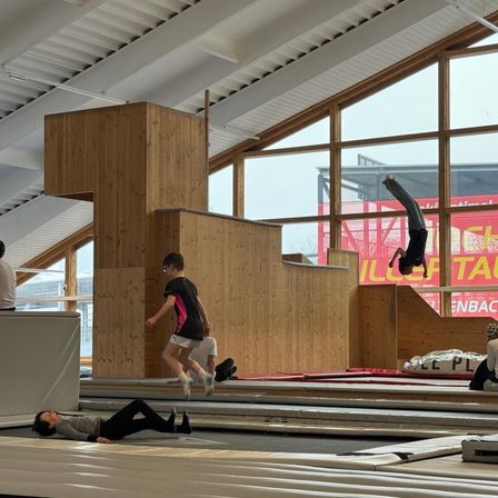 In einer Indoor-Einrichtung führt eine Person eine Trickaktion in der Luft auf einem Trampolin durch. Eine andere Person liegt auf dem Boden. Eine Person läuft die Holzstufen hinauf. Eine Person steht an der Seite. An der Wand befinden sich Banner.