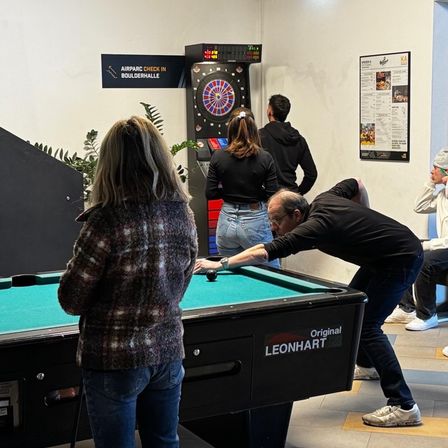 Eine Gruppe von Menschen spielt Darts in einem Raum mit einem grünen Tisch. Ein Mann beugt sich über den Tisch, und eine Frau steht hinter ihm. Das Dartboard hat eine digitale Anzeige.