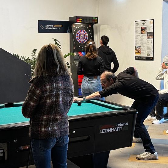 Eine Gruppe von Menschen spielt Darts in einem Raum mit einem grünen Tisch. Ein Mann beugt sich über den Tisch, und eine Frau steht hinter ihm. Das Dartboard hat eine digitale Anzeige.