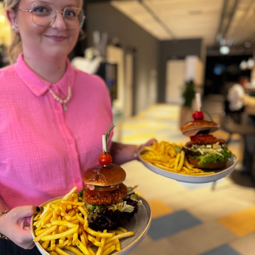 Eine Frau hält zwei Teller mit Burgern und Pommes, lächelt in einem Restaurant.