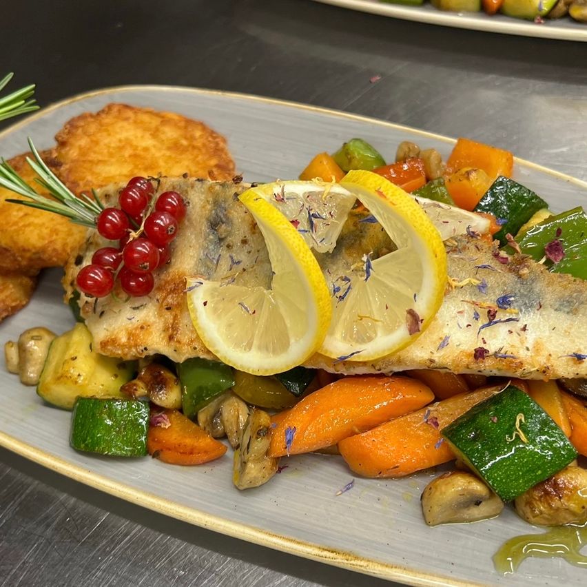 Ein Teller mit gegrilltem Fischfilet mit Zitronenscheiben, Kräutern und verschiedenen Gemüsesorten wie Karotten, Paprika und Pilzen.