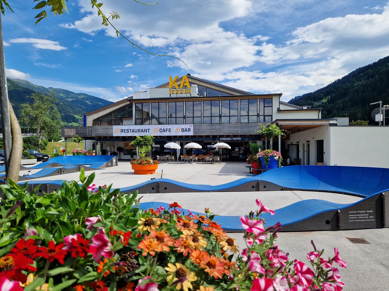 Ein modernes Gebäude mit dem Schild KA BOOM in gelben Buchstaben. Im Vordergrund befinden sich Pflanzen und Blumen sowie blaue Rampen. Es gibt Sonnenschirme, Stühle und Tische draußen. In der Ferne sind Berge zu sehen.