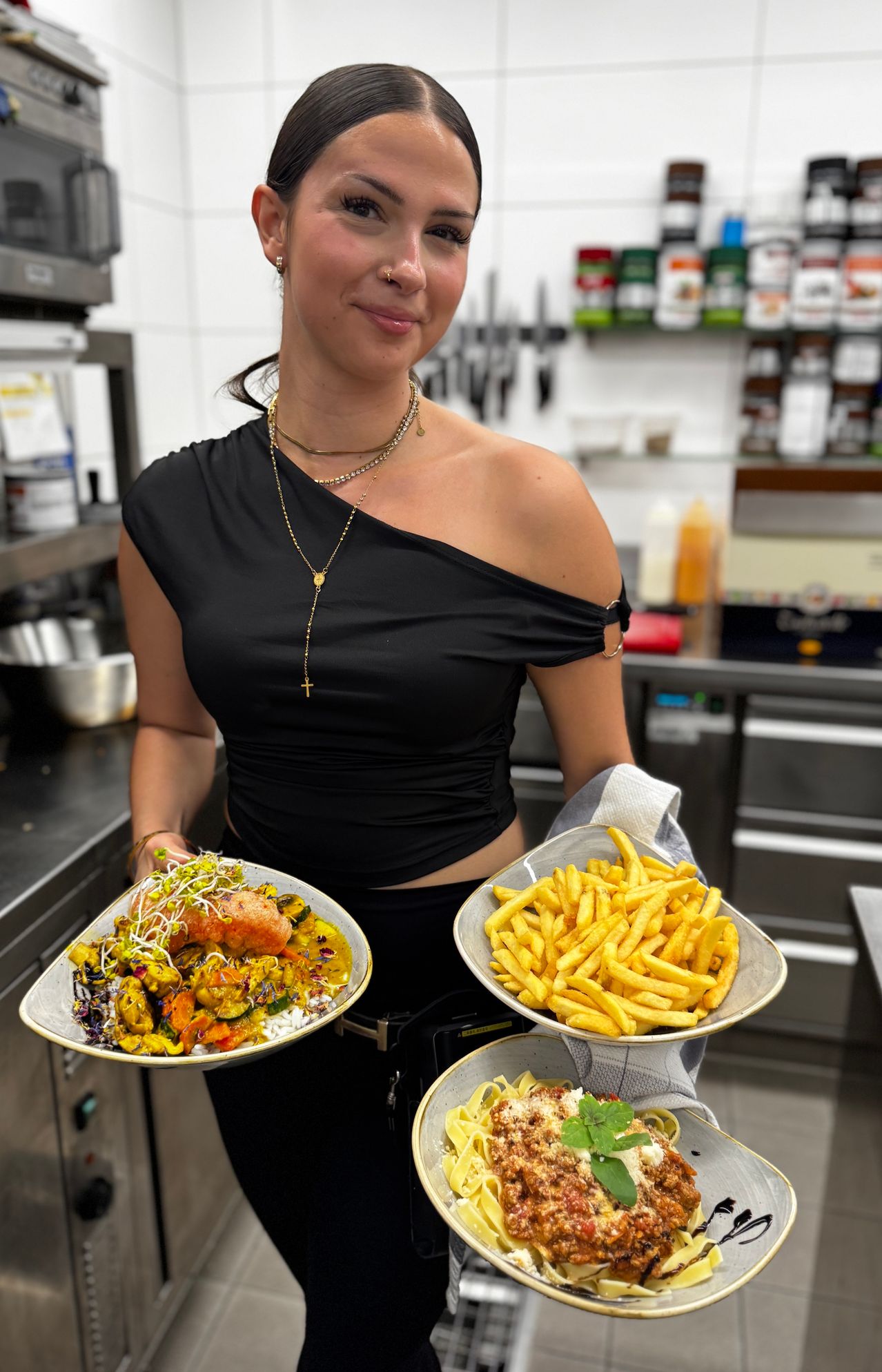 Eine Frau in einem schwarzen Oberteil hält zwei Teller mit Essen in einer Küche. Ein Teller hat ein Gericht mit Gemüse und Reis, der andere hat Pommes Frites.
