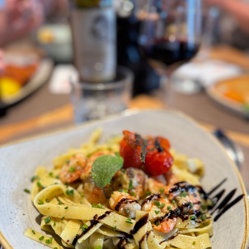 Ein Teller mit Pasta, Garnelen, Kirschtomaten und Basilikum-Garnitur auf einem Holztisch, begleitet von einem Glas Wein und einer Flasche Wein.